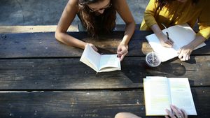 People read books and write in a notebook at a sunny wooden picnic table
