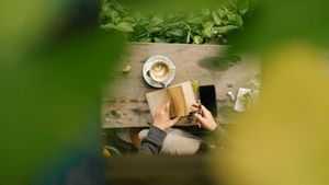 A person reading an open book beside a cup of coffee at a wooden table in morning light
