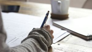 A student writing notes with a pen in an open paper journal beside a laptop on a sunny wooden desk