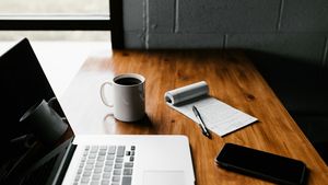 A clean workspace with an open notebook, laptop, pen, and coffee cup on a sunlit white desk