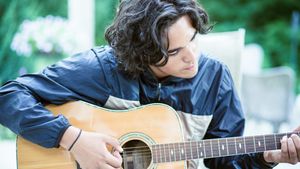 A young man with curly hair plays an acoustic guitar outside in a green garden