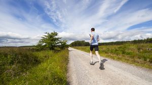A person jogging down a sunlit trail between green fields under a bright blue sky
