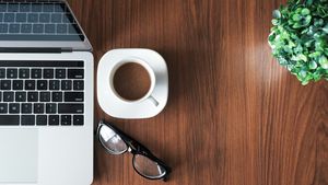 Overhead view of a laptop, white coffee cup, eyeglasses, and green plant on a sunlit wooden desk