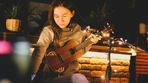 A woman sitting in a sunlit room practicing chords on a wooden ukulele