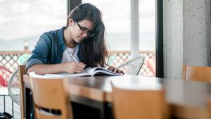 A woman in glasses and a denim shirt writes in a workbook at a table by a bright window