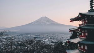 A red five-story pagoda stands in front of snow-capped Mount Fuji and a town at dusk
