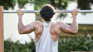 Man in a gray tank top performing a strict pull-up on an outdoor metal bar in warm sunlight