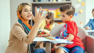 A teacher in an orange headscarf holds up her fingers to teach a young boy in a bright classroom